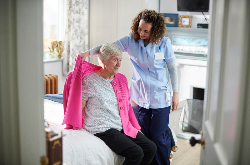 Respite Stay Caregiver helping a senior woman put on a jacket in a warmly furnished apartment at Greenleaf Court Senior Living